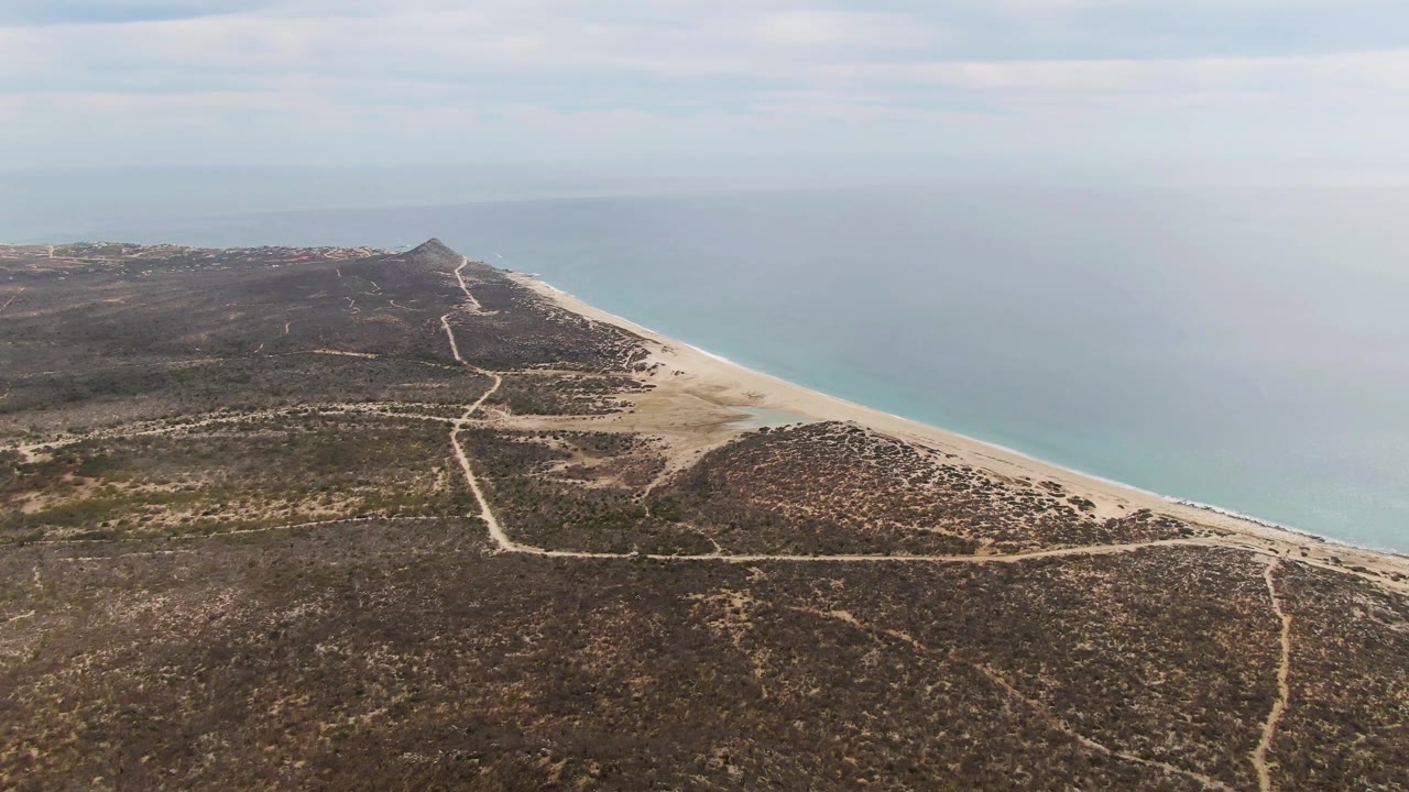 High aerial view at Punta Gorda Beach with branching dirt tracks across brown rocky land, a pale sand beach on the right, calm blue water beyond, and no visible people.