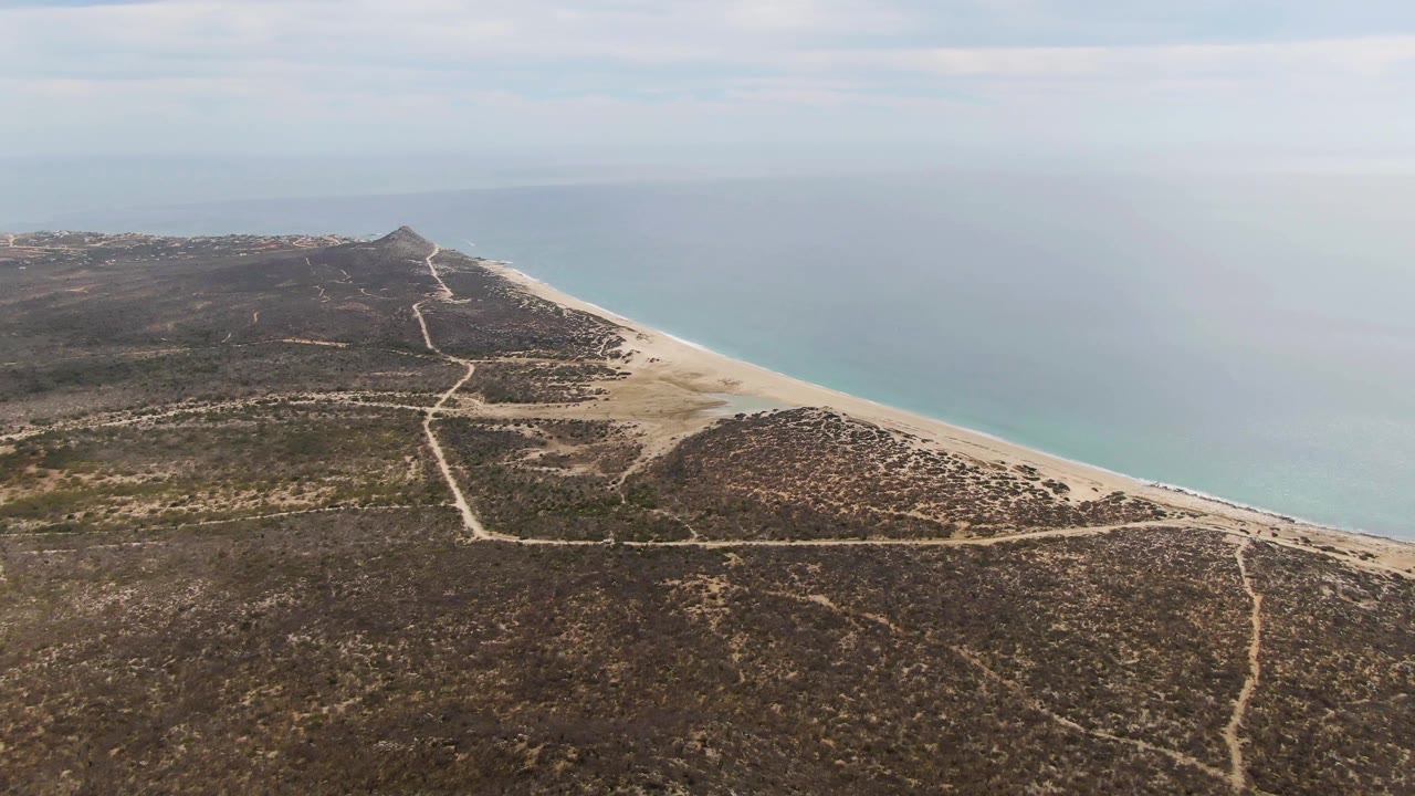 Aerial frame of Punta Gorda Beach showing a central dirt road through dry coastal hills, the sandy shore curving along the right edge, blue ocean, clouded horizon, and no visible people.