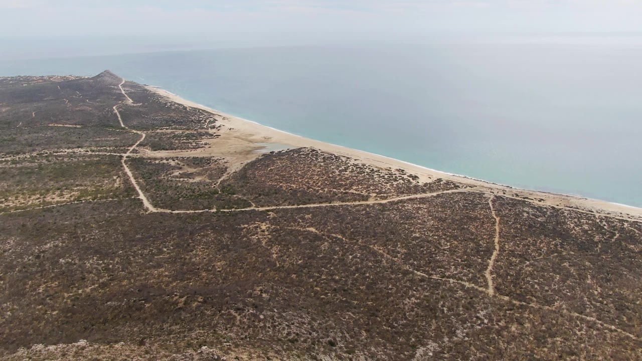 Second frame from Punta Gorda Beach shows the shoreline running diagonally beside calm blue water, with rocky scrubland and pale dirt tracks across the foreground, no visible people.