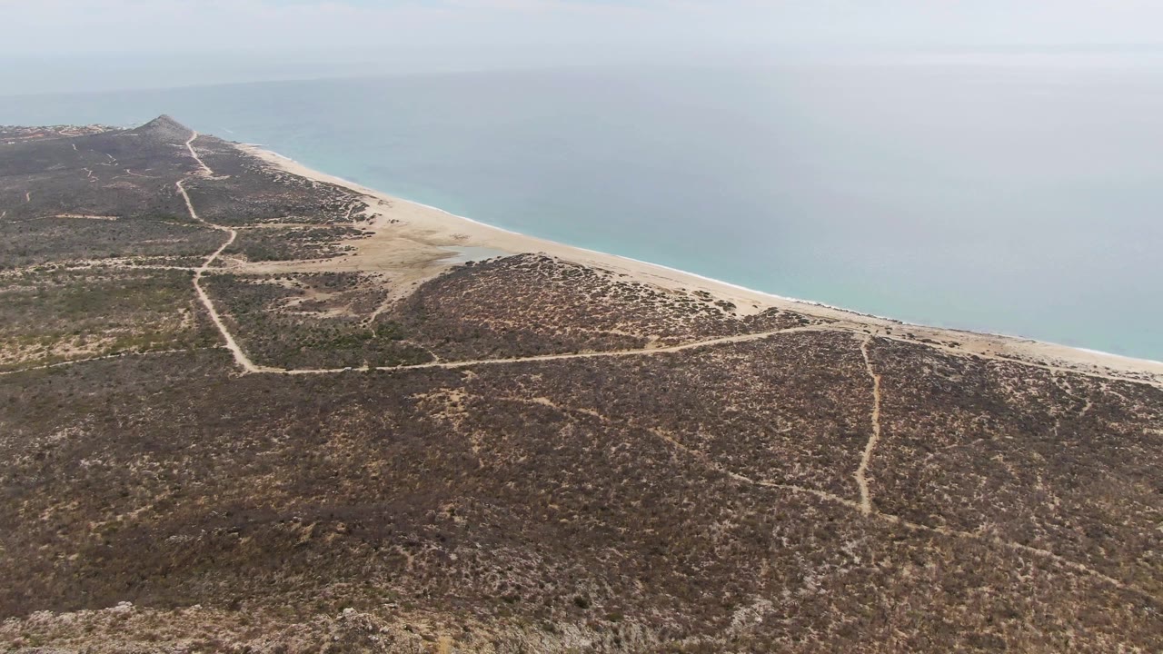 Aerial view at Punta Gorda Beach with the headland farther along the coast, pale sand between scrubland and ocean, visible dirt paths, and no visible people.
