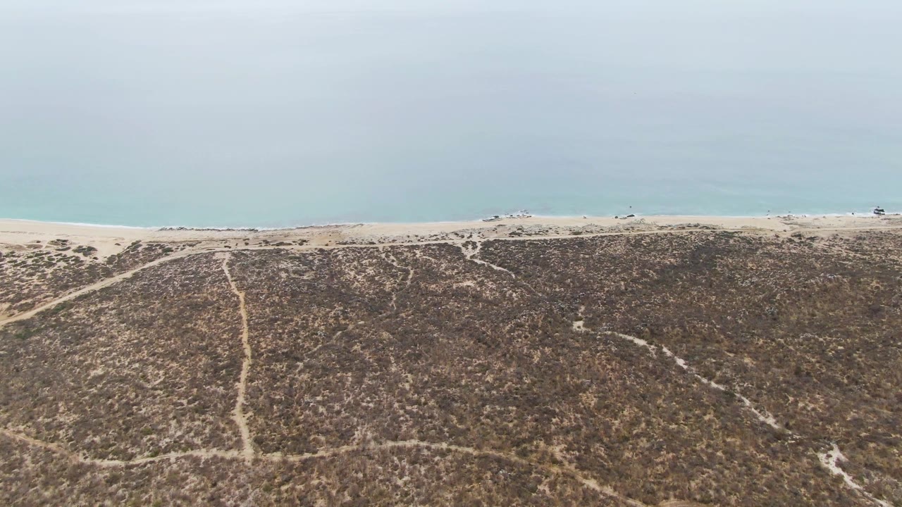 Aerial frame at Punta Gorda Beach showing scrub-covered dunes with faint paths, a narrow sand strip, pale ocean water, and tiny distant beach activity.