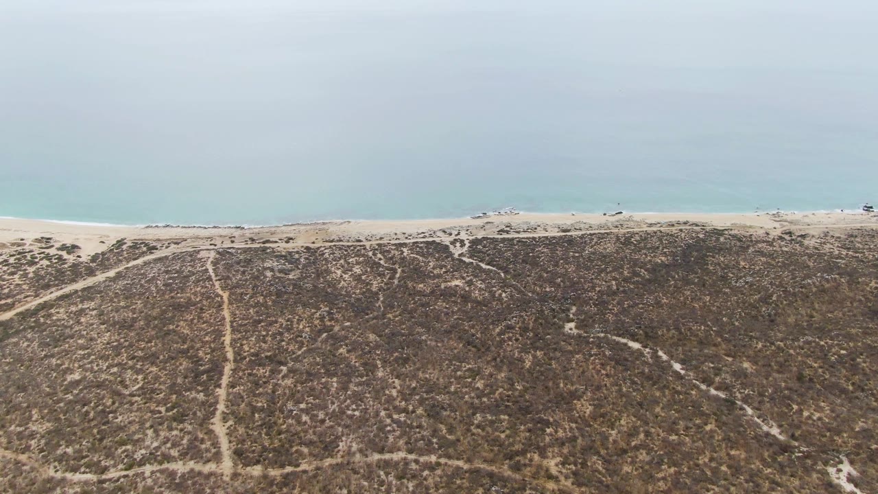 High view over Punta Gorda Beach with diagonal dune tracks, scrub vegetation, a pale sand shoreline, calm blue-green water, and sparse distant beach activity.