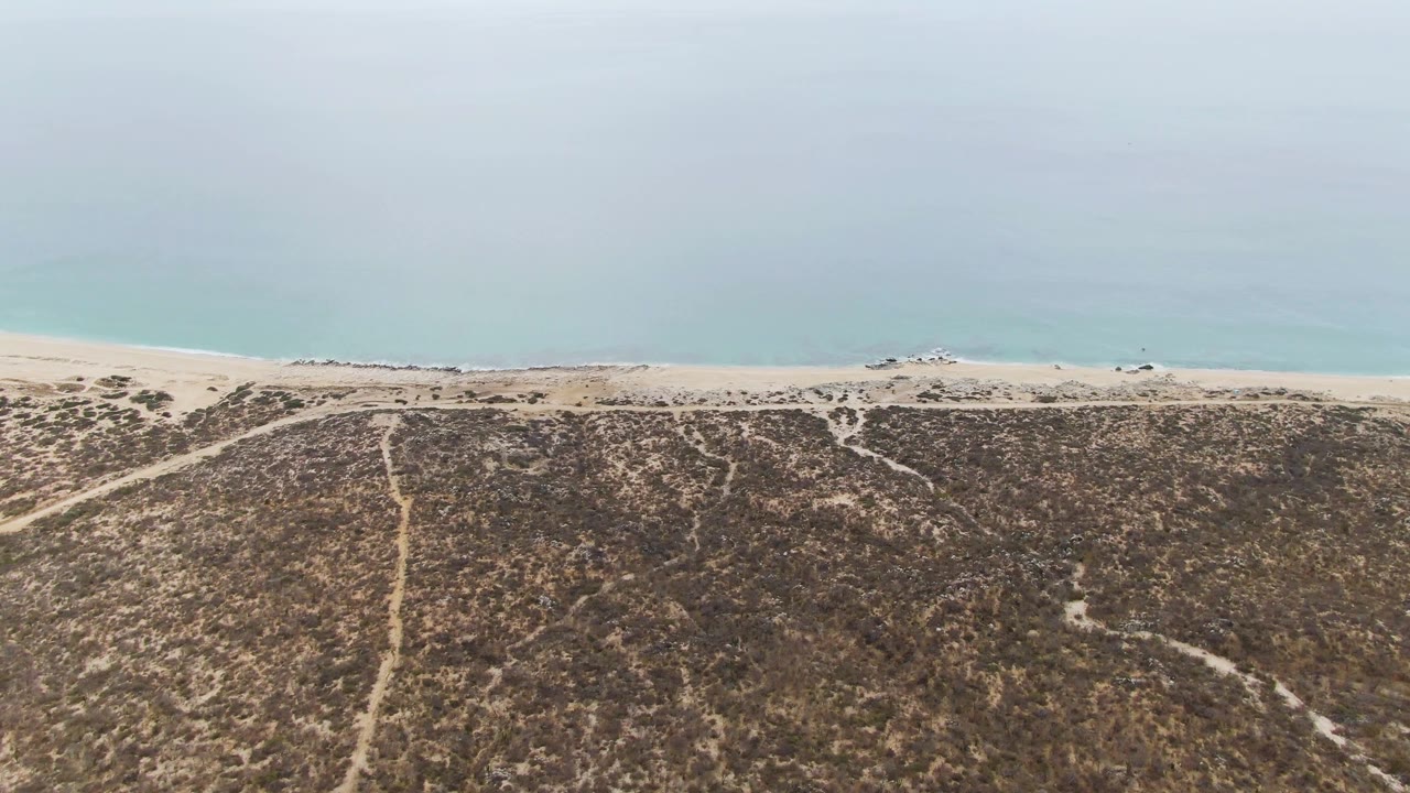 Mid-clip aerial frame from Punta Gorda Beach with dry vegetation covering the dunes, worn sand paths leading to the beach, pale blue water beyond, a small vehicle at the shoreline, and no visible people.