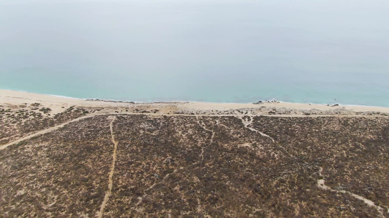 Aerial frame over Punta Gorda Beach, Baja California Sur, with scrub-covered dunes in the foreground, pale sand at the shore, and calm blue water under overcast light.