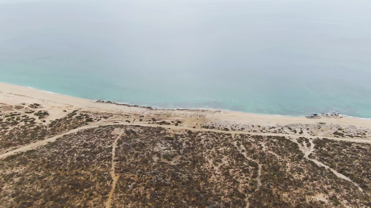 Aerial coastline with sandy tracks through dunes and light blue water beyond.