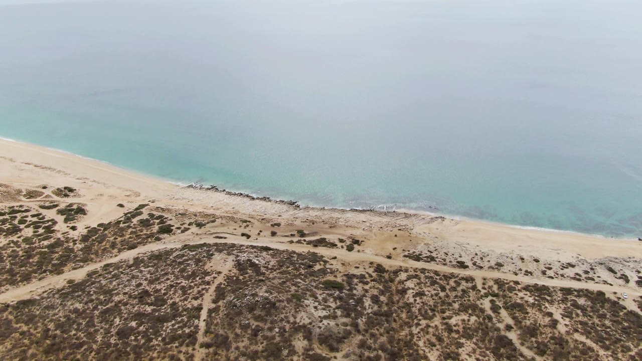 Aerial frame at Punta Gorda Beach with turquoise water across the top, pale sand through the center, scrubby dunes below, and no visible people.