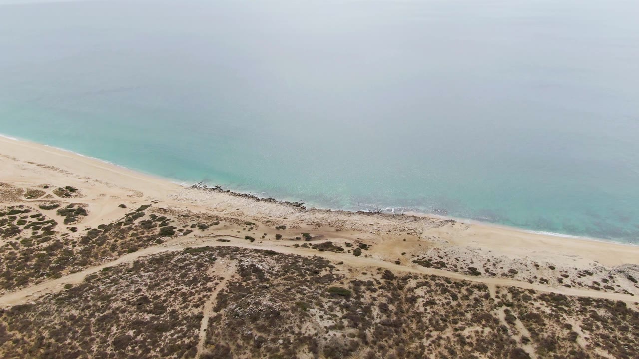 Aerial coastline frame from Punta Gorda Beach with the surf line slightly farther along the sand, scrub dunes in the foreground, and no visible people.