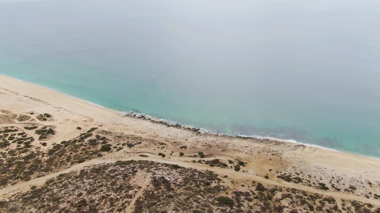 Aerial frame at Punta Gorda Beach showing a thin white surf edge against sand, muted turquoise water, low scrub on dunes, and no visible people.