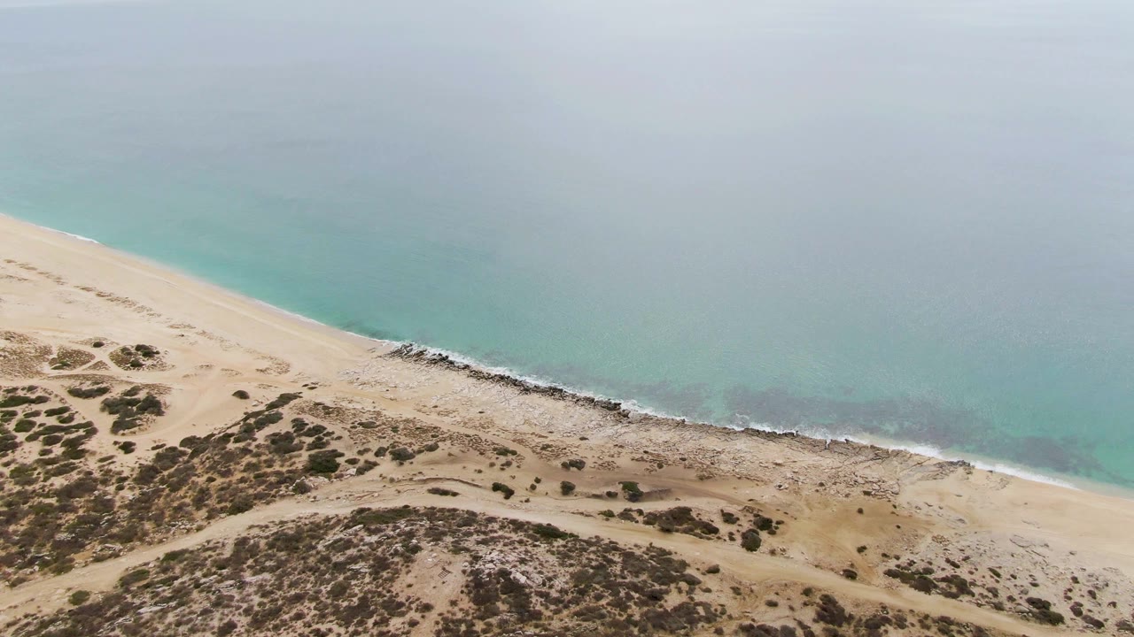 Mid-clip aerial frame over Punta Gorda Beach showing calm turquoise water, a narrow foam line on the sand, a faint dune track, sparse vegetation, and no visible people.