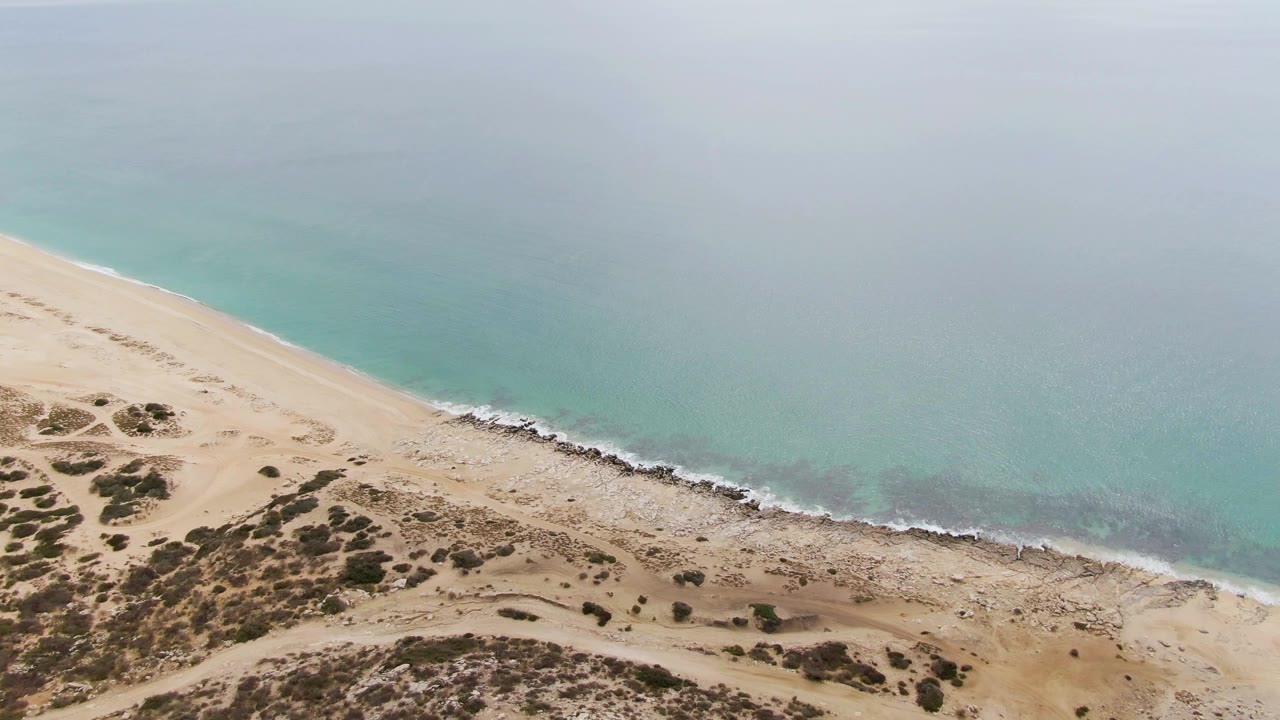 Aerial view over Punta Gorda Beach in Baja California Sur, with sandy dunes and sparse scrub below a diagonal rocky shoreline and turquoise water, no visible people.