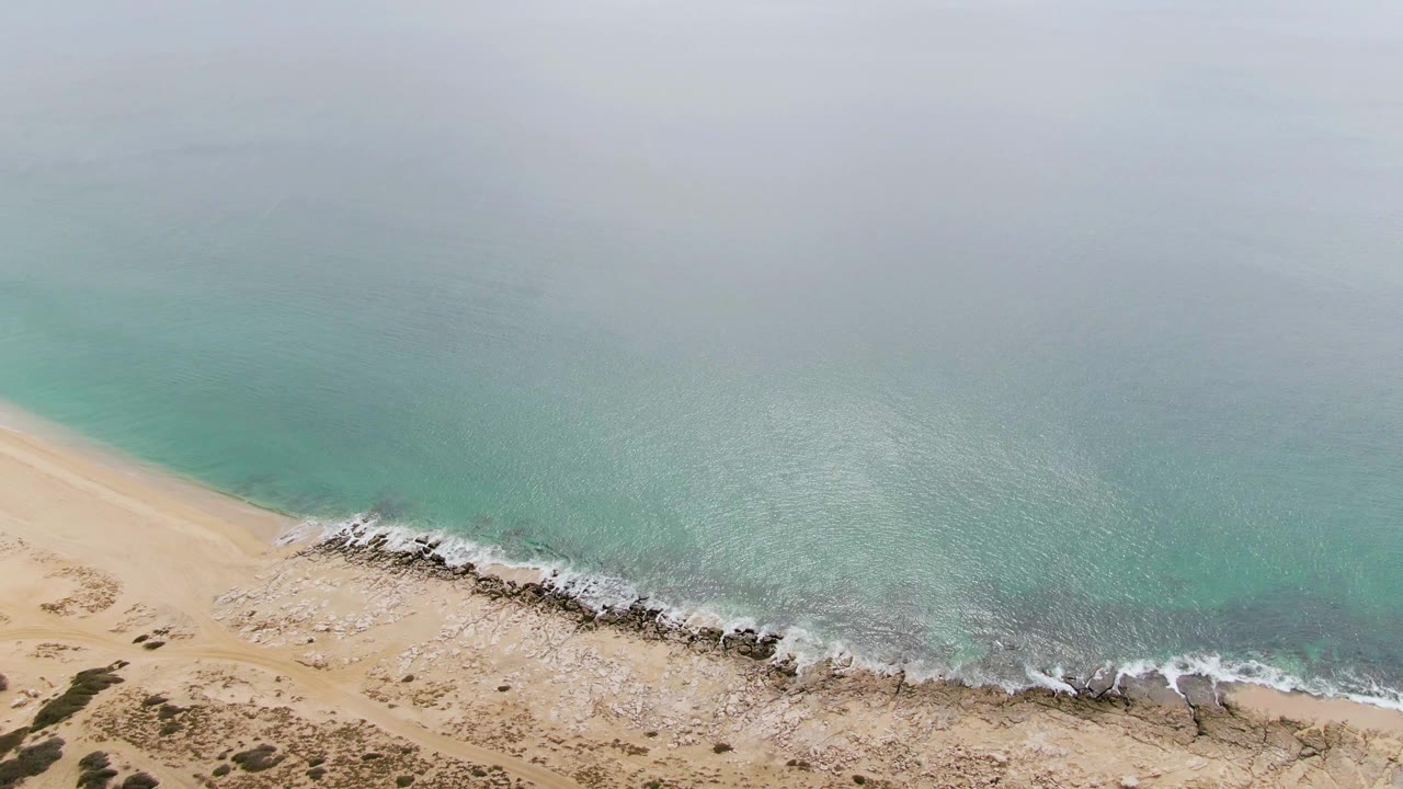 Aerial view of Punta Gorda Beach with the surf line shifted along the rocky edge, pale sand at lower left, open turquoise water above, and no visible people.