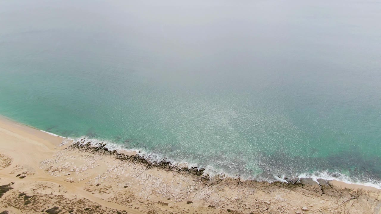 High aerial frame at Punta Gorda Beach showing a longer run of dark shoreline rocks, white wave foam, tan sand, muted turquoise sea, and no visible people.