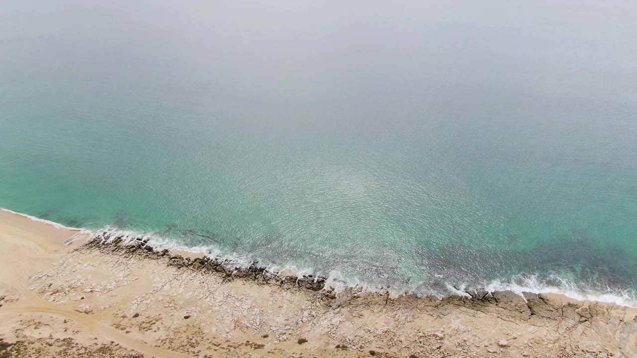 Aerial view over Punta Gorda Beach with more open water filling the upper frame, foam washing across a dark rocky strip, pale sand below, and no visible people.