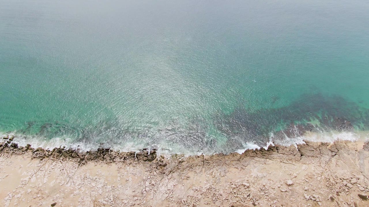 Aerial frame over Punta Gorda Beach with a thin white wave line crossing dark rocks and pale sand, no visible people.