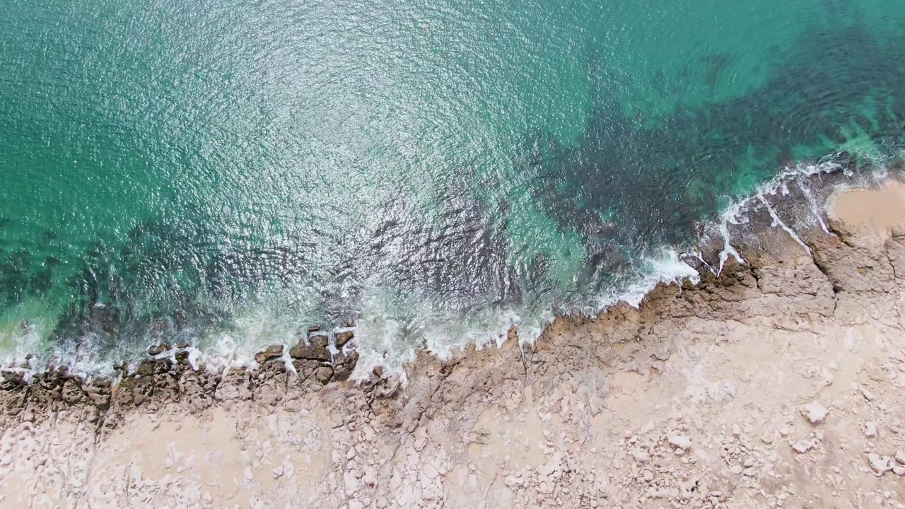Top-down view of Punta Gorda Beach turquoise water brushing a pale rocky shoreline, with darker submerged rocks offshore and no visible people.