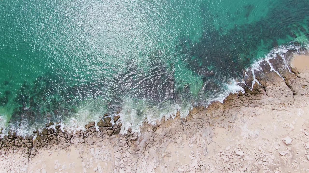 Aerial frame at Punta Gorda Beach showing a slightly broader band of foam over tan rocks as clear green-blue water reaches the shoreline, with no visible people.
