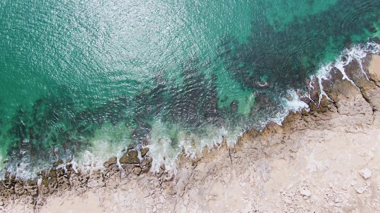 Overhead Punta Gorda Beach view with turquoise water receding from jagged pale rocks, leaving thin white foam along the diagonal shore and no visible people.