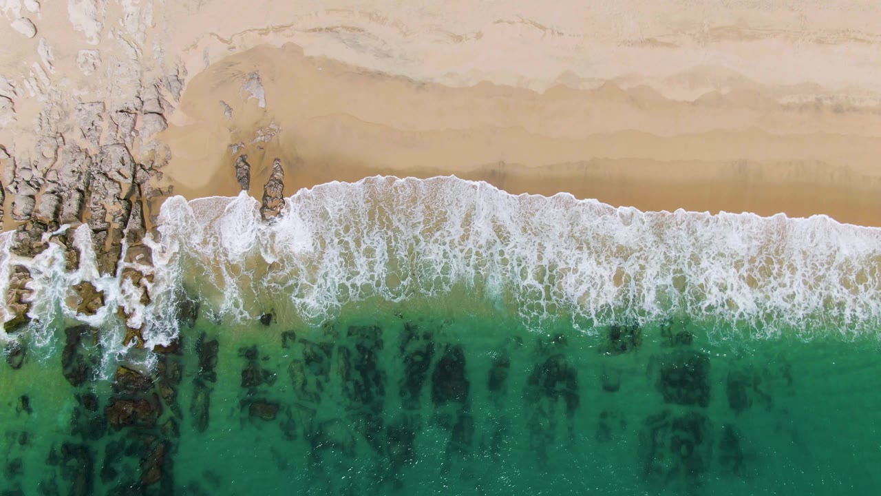 Overhead view of Punta Gorda Beach in Baja California Sur, with white foam breaking across pale sand and turquoise shallows over dark rock patches, no visible people.