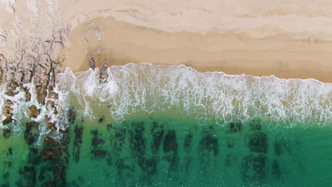 Aerial frame over Punta Gorda Beach where the wave edge pushes toward the sand beside a band of rocky outcrops and green-blue water, no visible people.