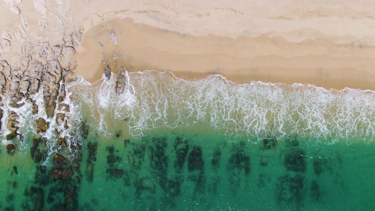 Top-down Punta Gorda Beach frame with whitewater spread wider across the shore, tan sand above, and turquoise water with dark reef-like rock shapes below, no visible people.