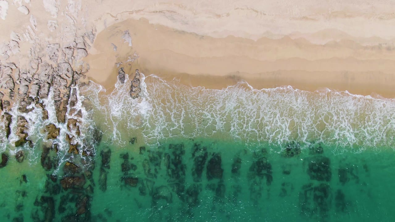 Overhead beach frame with rocks at left and foamy waves over turquoise shallows.