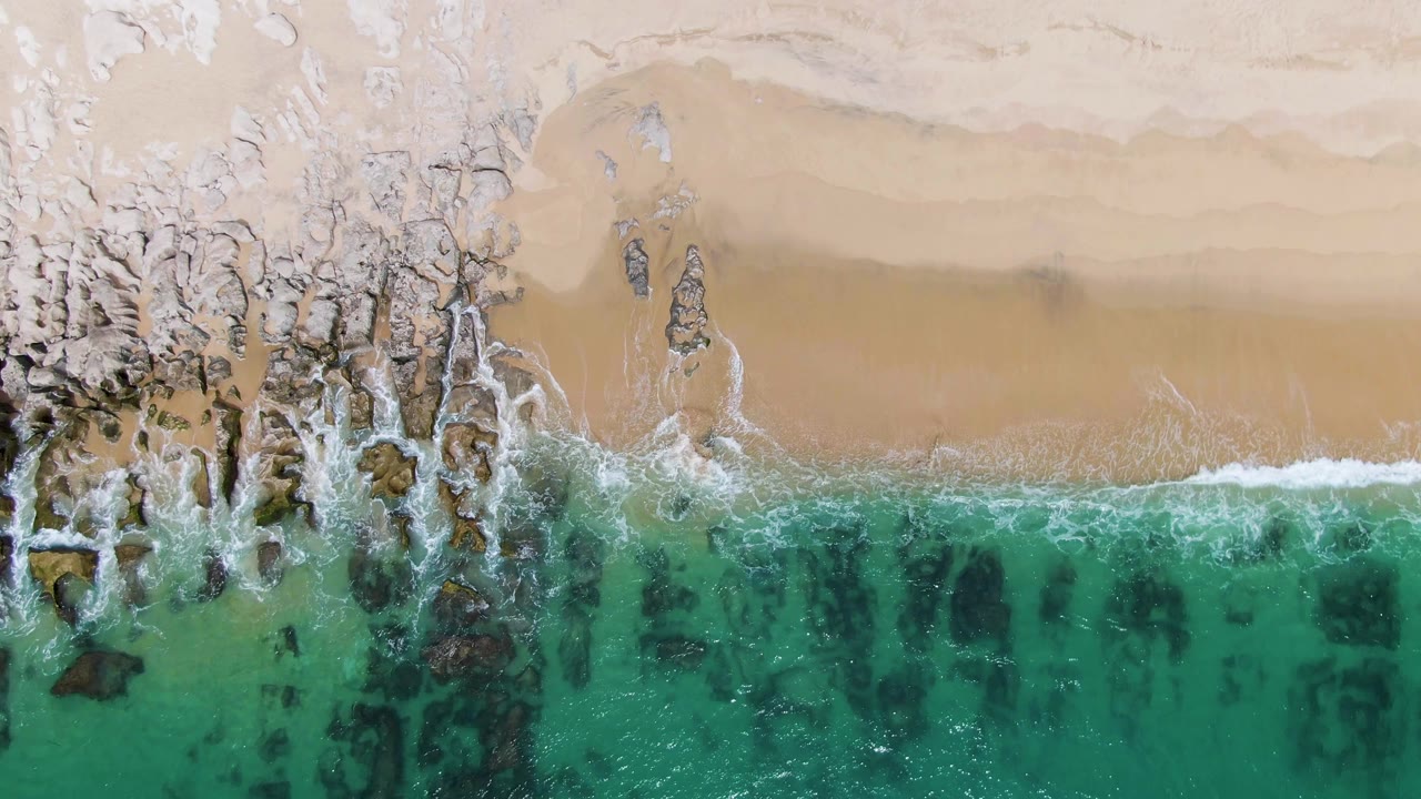 Top-down aerial frame of Punta Gorda Beach in Baja California Sur, Mexico, showing turquoise water, dark rocks, pale sand, and a thin white surf line with no visible people.