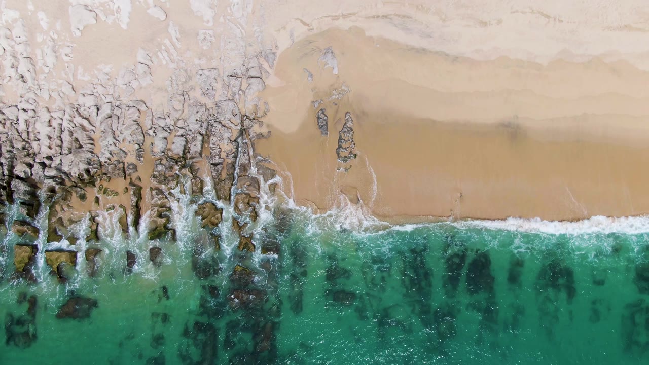 Aerial view at Punta Gorda Beach with white foam widening over clustered dark rocks as turquoise water presses toward the sandy shore, with no visible people.