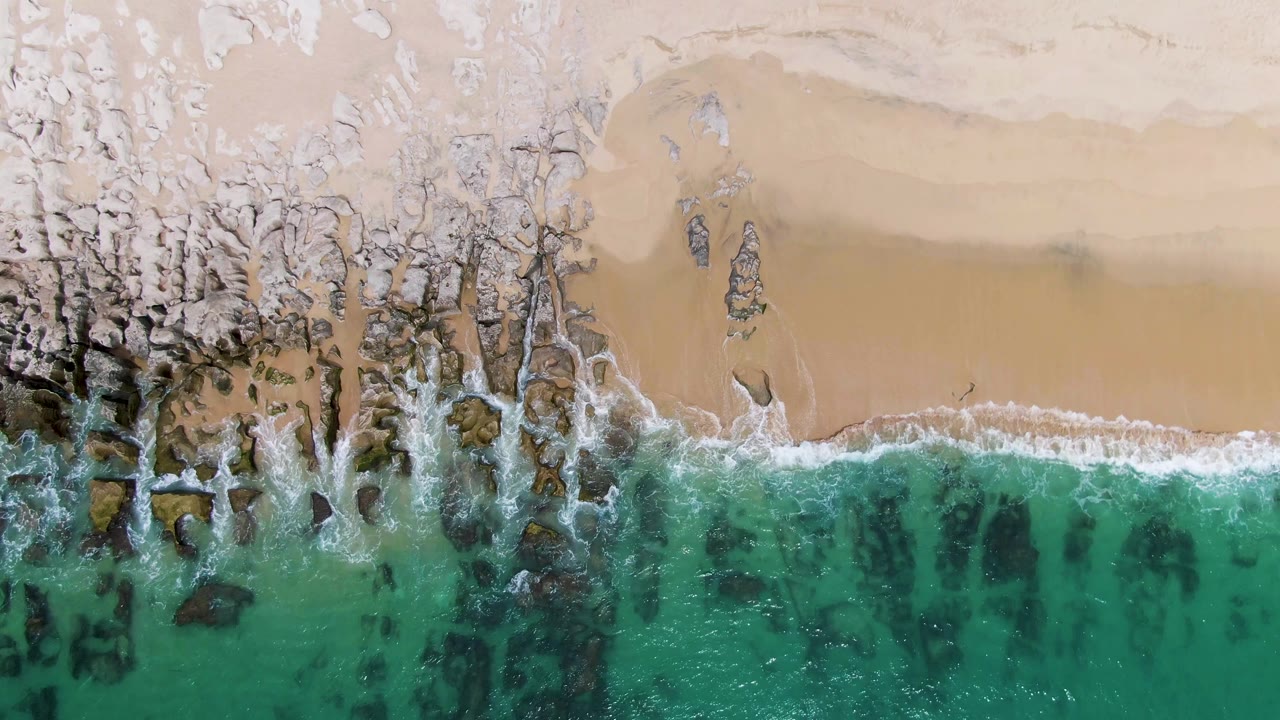 Top-down Punta Gorda Beach frame showing a curved white wave edge along pale sand, dark rock clusters near the break, and green-turquoise water below with no visible people.
