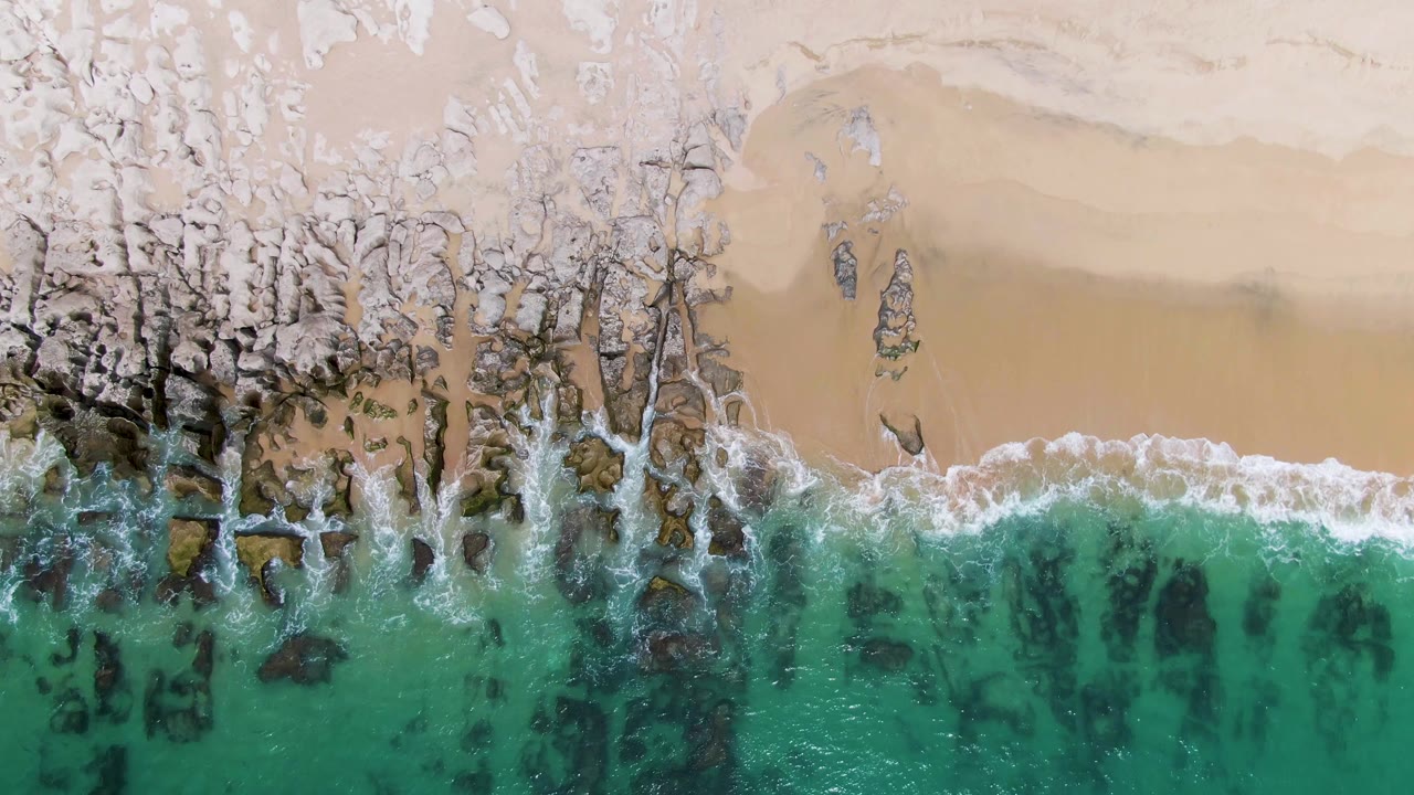 Aerial shoreline with foam retreating around exposed rocks and sand.