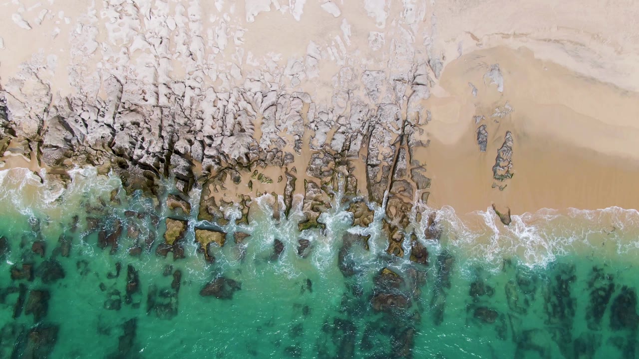 Overhead frame at Punta Gorda Beach, Baja California Sur, with turquoise water reaching a pale sandy shore through dark exposed rocks and no visible people.
