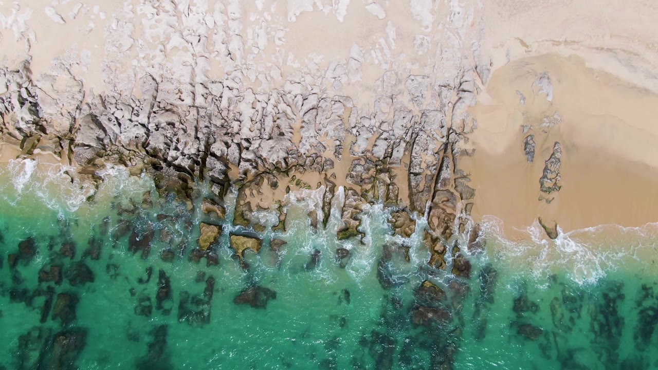A later overhead frame at Punta Gorda Beach shows white foam shifting along the central rock band between turquoise water and pale sand, with no visible people.