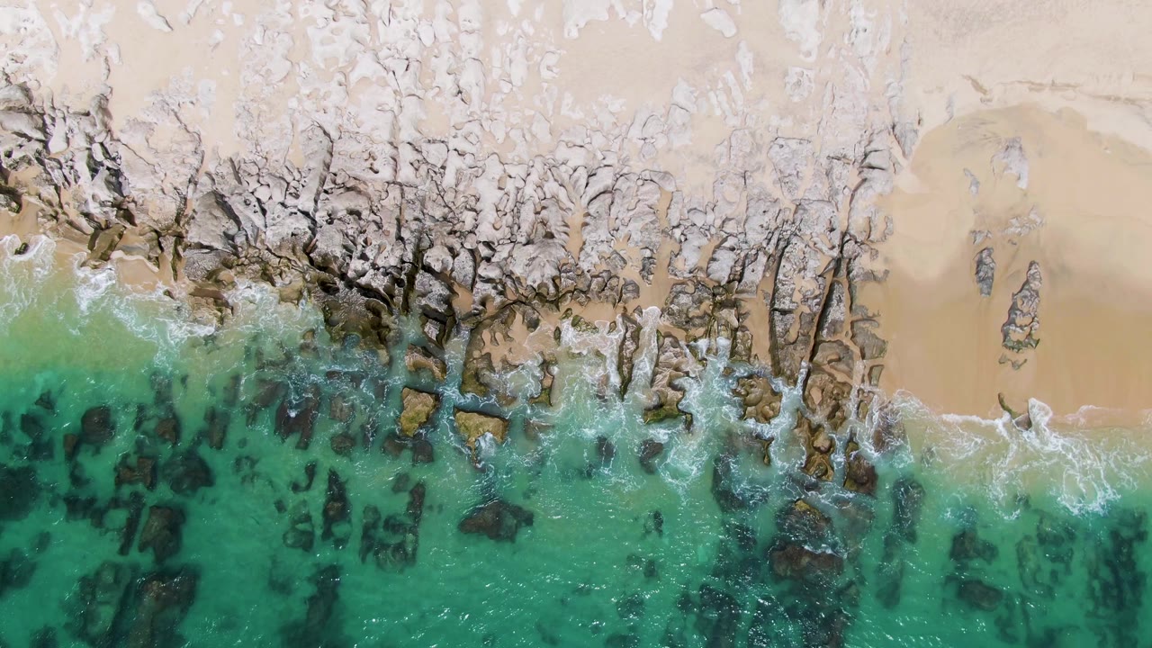 At 0:02, the Punta Gorda Beach shoreline remains framed from above, with clear green-blue water below a jagged rock edge and light sand above, and no visible people.