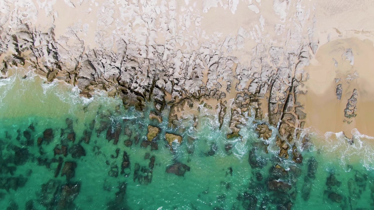 Overhead shoreline with dark submerged rocks, white surf, and pale sand.