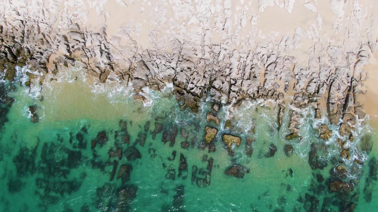 Overhead view of Punta Gorda Beach with turquoise water over dark submerged rocks, pale rocky shore, tan sand, and no visible people.