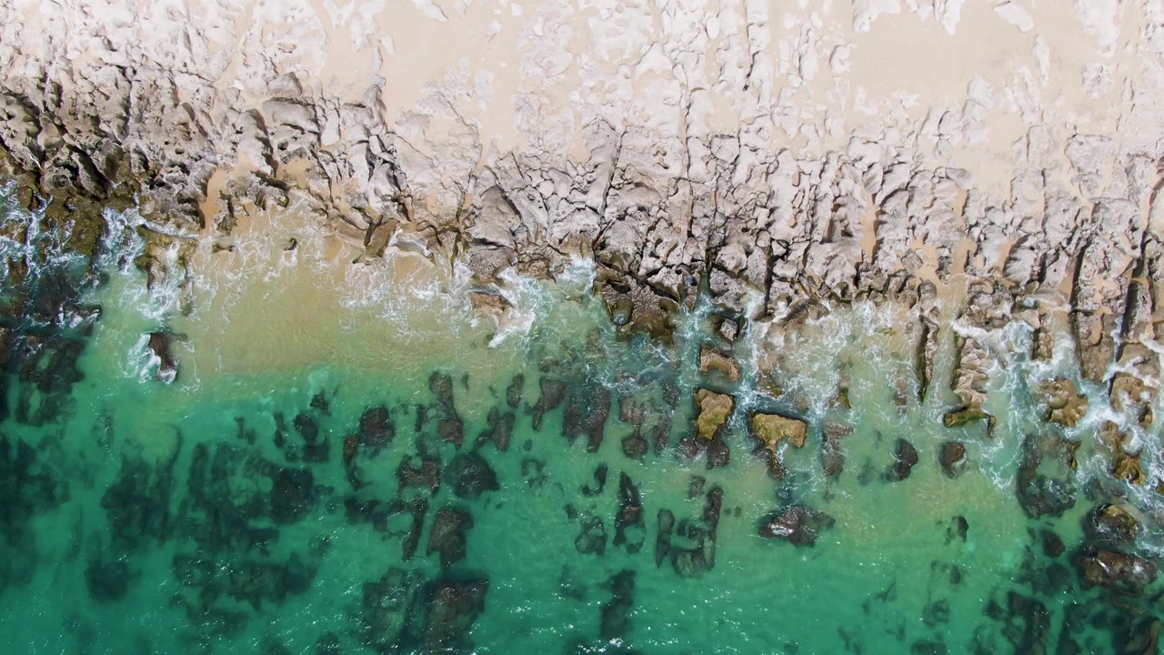 Punta Gorda Beach shoreline from above with a thin foam line along the sand, scattered dark rocks under green turquoise water, and no visible people.
