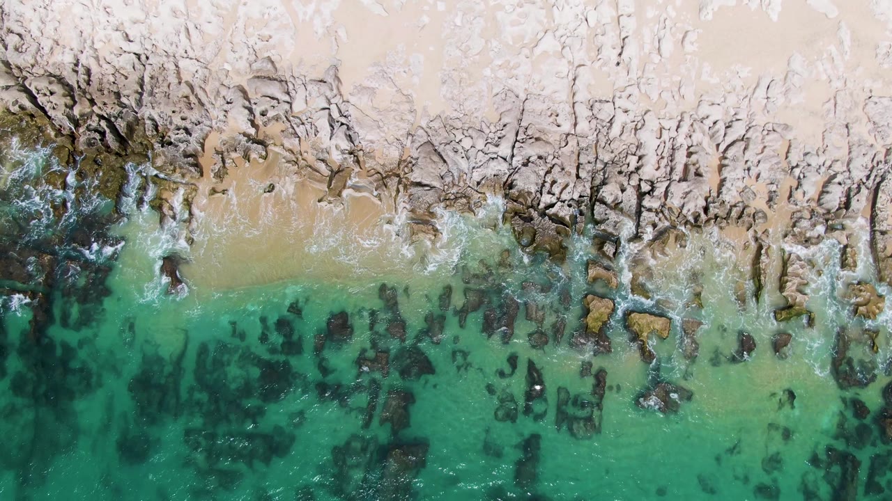 Straight-down view of Punta Gorda Beach where clear shallow water reveals dark rock patches below a pale rocky and sandy shoreline with no visible people.