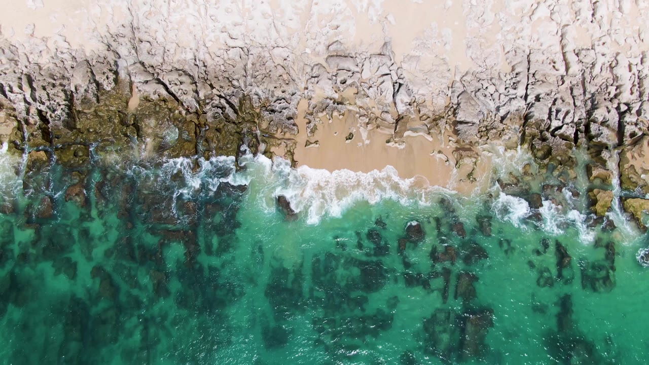 Aerial frame at Punta Gorda Beach showing white foam pushing farther over the rocky sand edge while clear turquoise water reveals dark rocks below, no visible people.