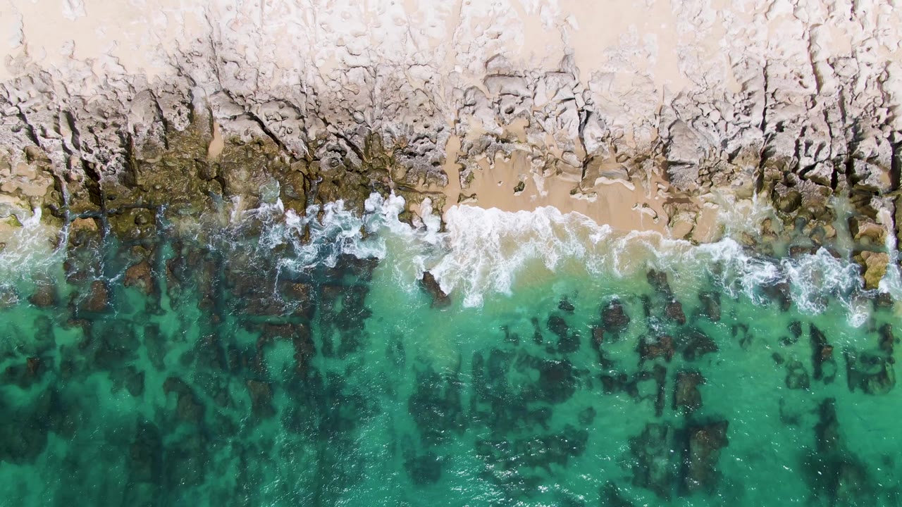 Top-down Punta Gorda Beach frame with a wider white surf band breaking across the rocky shoreline and mottled dark rocks under the green-blue water, no visible people.