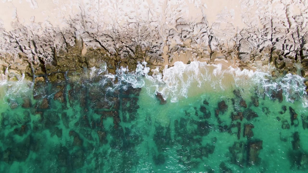 Overhead view of Punta Gorda Beach with the foam line receding unevenly across rocks, pale sand above, and transparent turquoise water below, no visible people.
