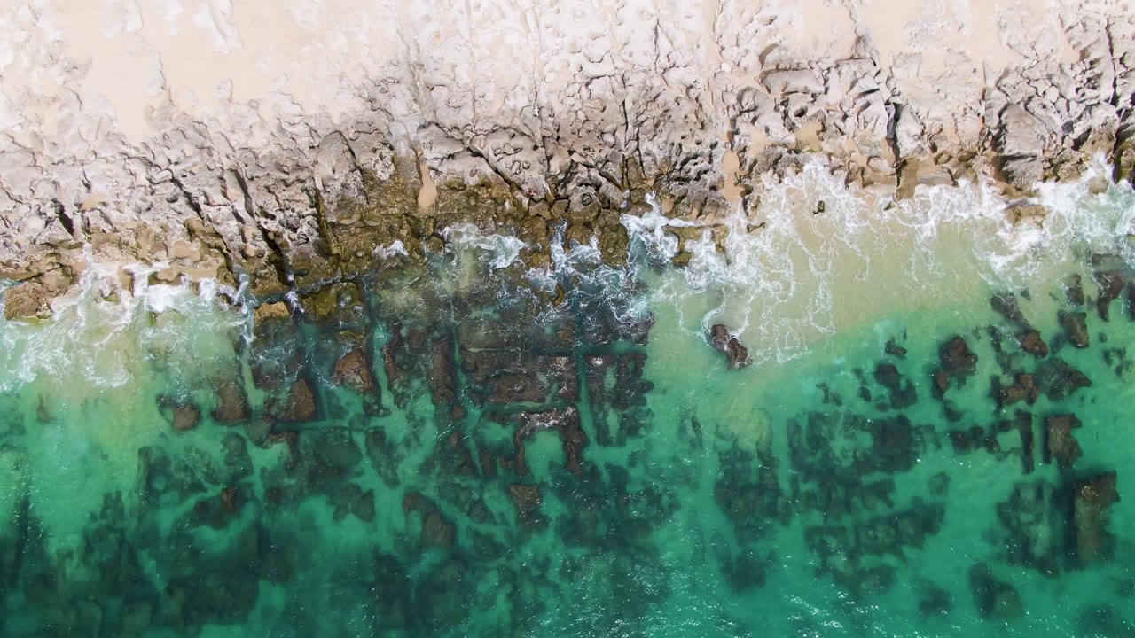 Top-down aerial frame at Punta Gorda Beach, Baja California Sur, showing pale sand, dark rock formations, thin wave foam, and clear turquoise water with no visible people.