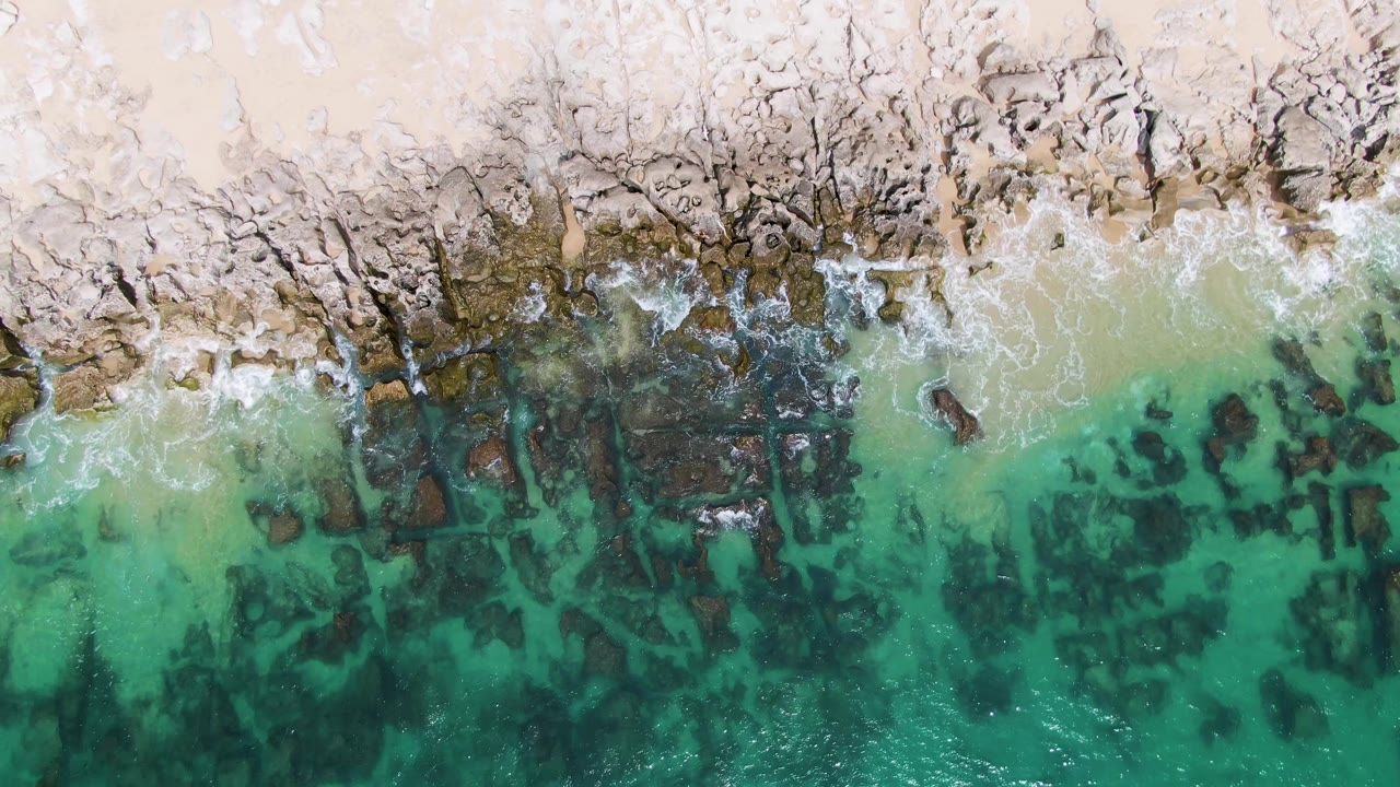 Aerial shoreline with white surf brushing rocks and clear green water.