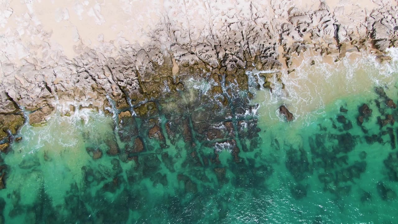 At 0:02, the Punta Gorda Beach shoreline remains in overhead view, with small waves breaking over dark rocks and pale sand bordering the shallow turquoise water, no visible people.
