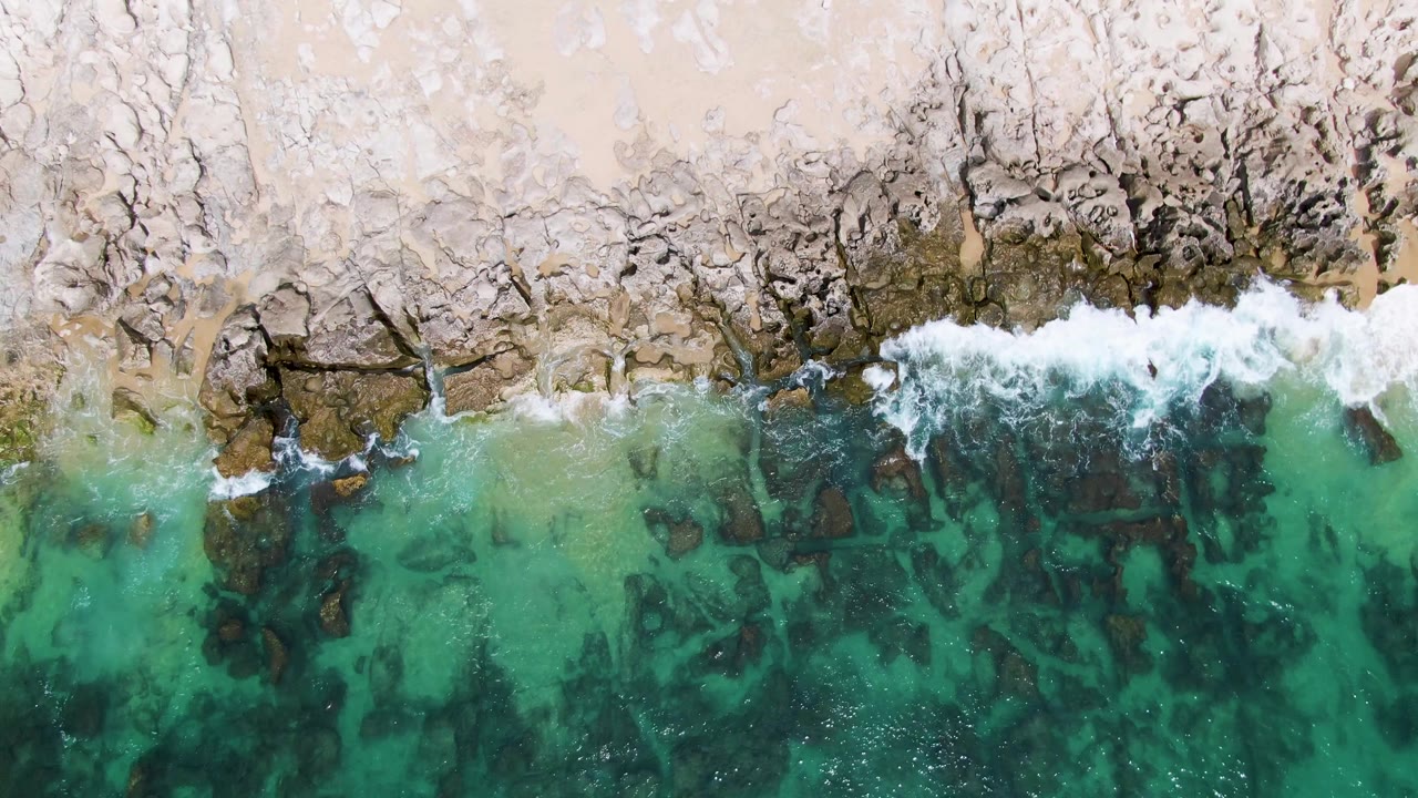 Aerial frame from Punta Gorda Beach, Mexico, with white foam spreading over dark rocks between pale sand and green-blue shallow water, with no visible people.