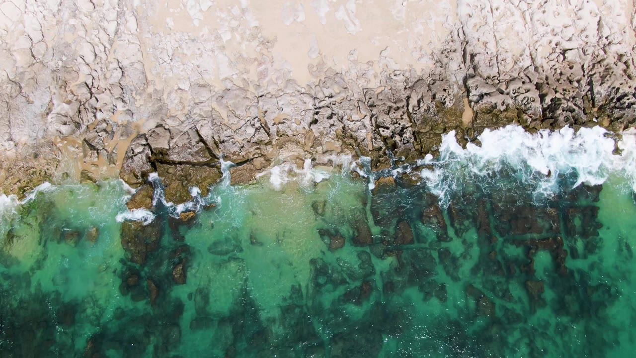 Top-down Punta Gorda Beach shoreline frame showing smaller foam patches along exposed rocks, pale sand at the top, and clear turquoise shallows below, with no visible people.