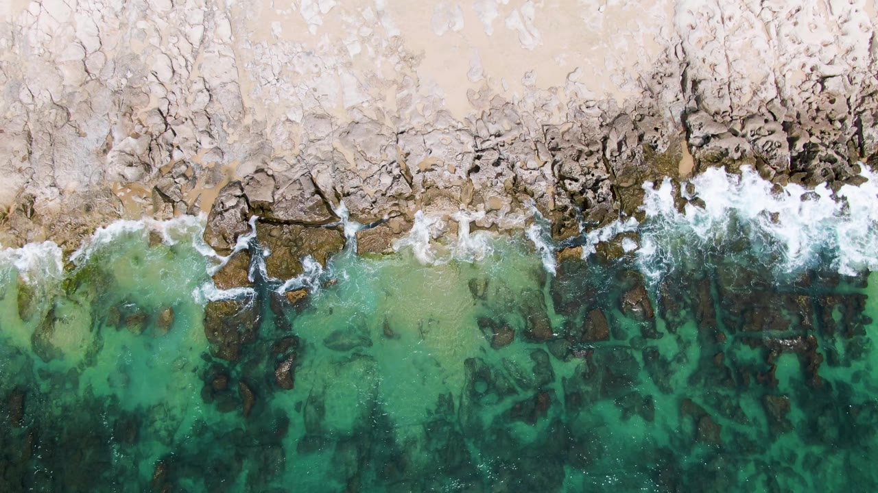 Overhead view at Punta Gorda Beach, Mexico, with surf foam washing over a darker rock cluster near the sand and green water filling the lower frame, with no visible people.