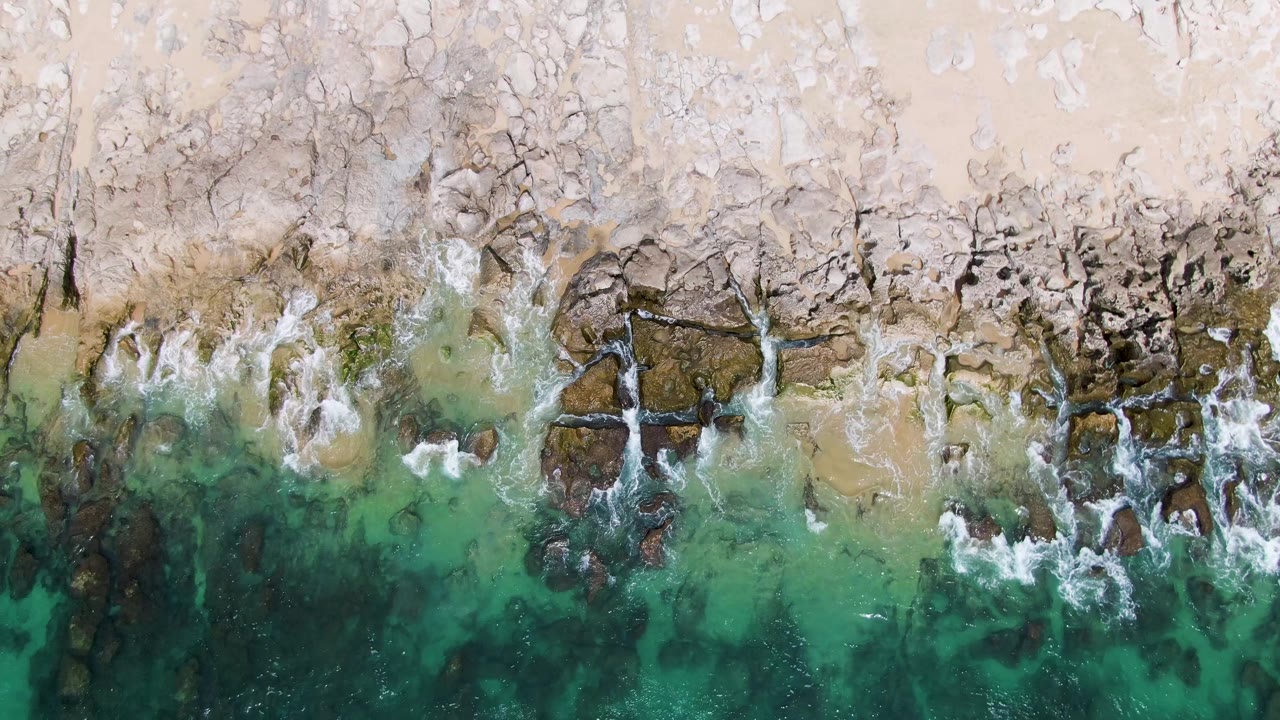 At 0:02, the aerial view of Punta Gorda Beach shows fractured pale rocks, darker tide-washed stone, and foamy waves crossing into clear water with no visible people.
