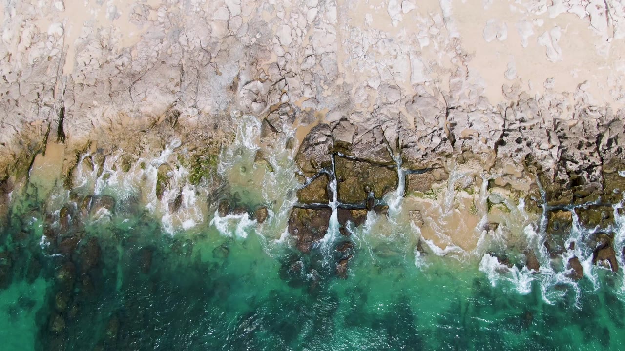 At 0:03, whitewater curls along the pale rocky edge of Punta Gorda Beach while deeper green water fills the lower frame and no people are visible.