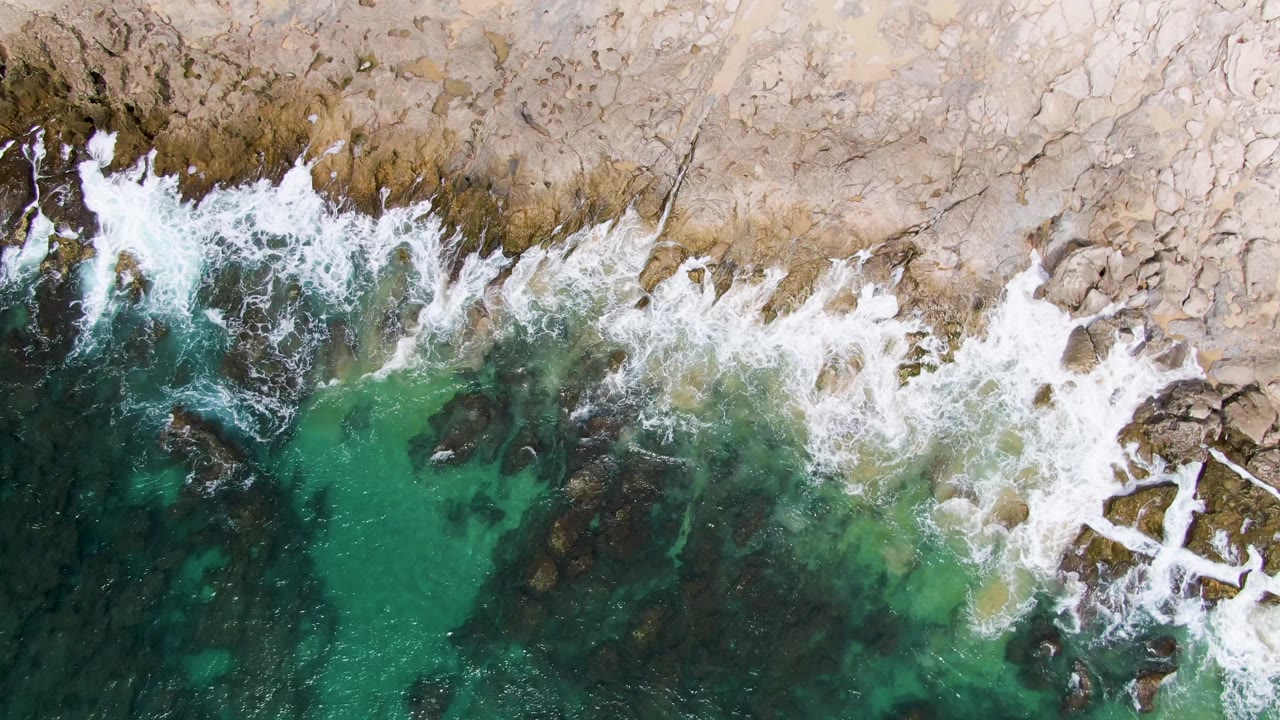 Overhead surf breaking on tan rocks beside turquoise water.