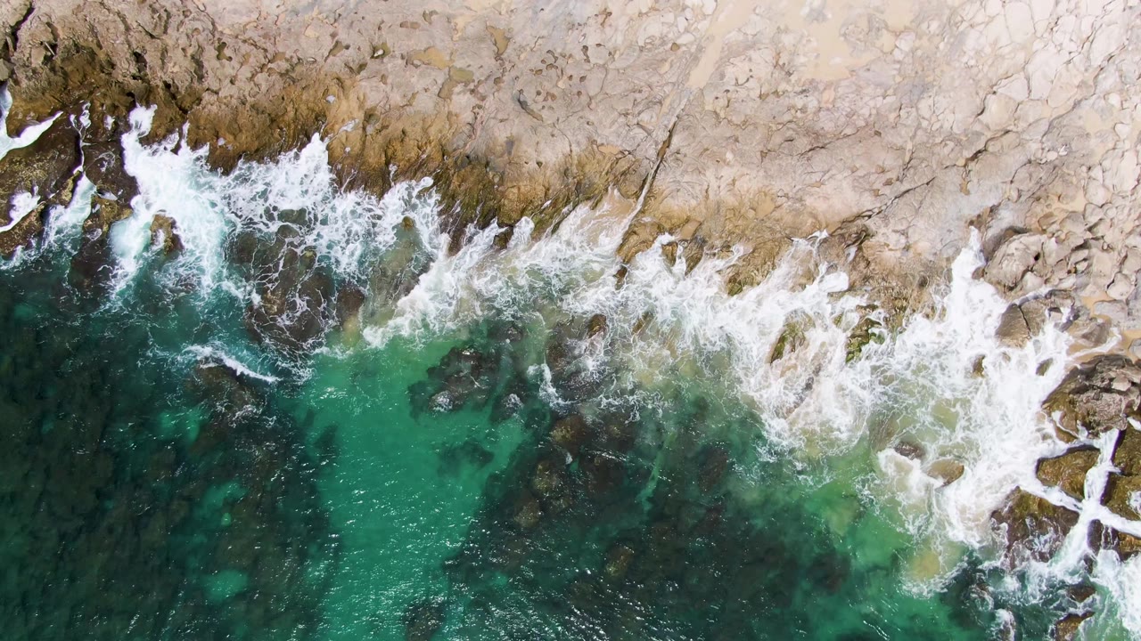 At Punta Gorda Beach in Baja California Sur, a larger line of white foam curls over the rock shelf while turquoise water and dark underwater stones remain visible below, with no visible people.