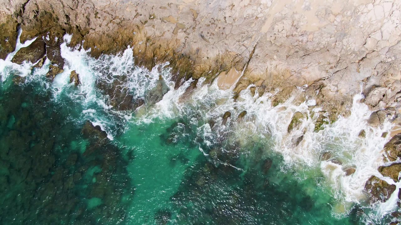 At Punta Gorda Beach in Baja California Sur, the surf line thins into streaks of foam along the upper rocks while green-blue water opens across the lower center, with no visible people.